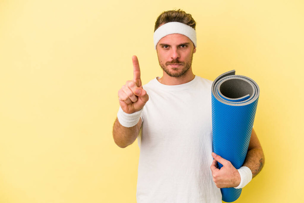Jovem esporte caucasiano homem indo para aulas de ioga enquanto segurando um tapete isolado no fundo amarelo mostrando o número um com o dedo. - Foto, Imagem