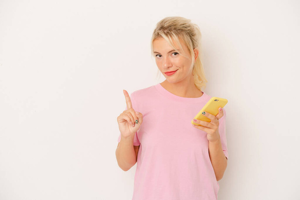 Young Russian woman holding mobile phone isolated on white background showing number one with finger. - 写真・画像