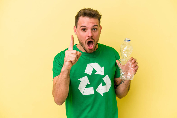 Młody biały mężczyzna trzymając butelkę plastiku do recyklingu izolowane na żółtym tle mając pomysł, pomysł inspiracji. - Zdjęcie, obraz