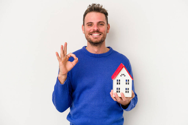 Young caucasian man holding a little house isolated on white background cheerful and confident showing ok gesture. - Фото, изображение