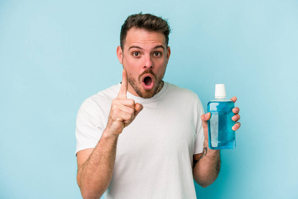 Young caucasian man holding mouthwash isolated on blue background having an idea, inspiration concept. - Foto, imagen