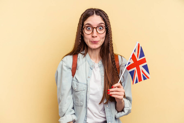 Young caucasian woman studying English isolated on yellow background shrugs shoulders and open eyes confused. - Photo, Image
