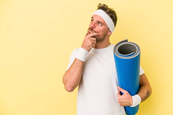 Young sport caucasian man going to yoga classes while holding a mat isolated on yellow background looking sideways with doubtful and skeptical expression. - Photo, Image