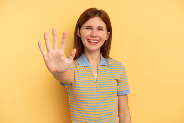 Sarı arka planda izole edilmiş genç İngiliz kadın gülümseyerek beş numarayı parmaklarıyla gösteriyor.. - Fotoğraf, Görsel