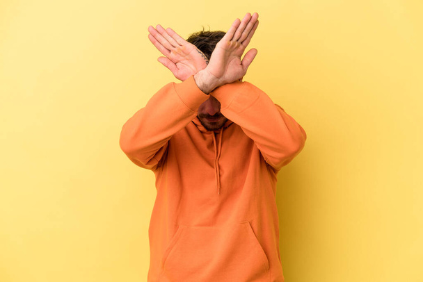 Молодой кавказский человек, изолированный на желтом фоне, держащий две руки скрещенными, концепция отрицания. - Фото, изображение