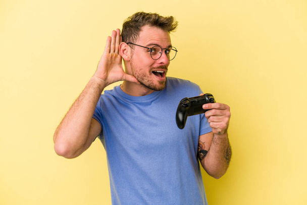 Giovane uomo caucasico in possesso di un controller di gioco isolato su sfondo giallo cercando di ascoltare un pettegolezzo. - Foto, immagini