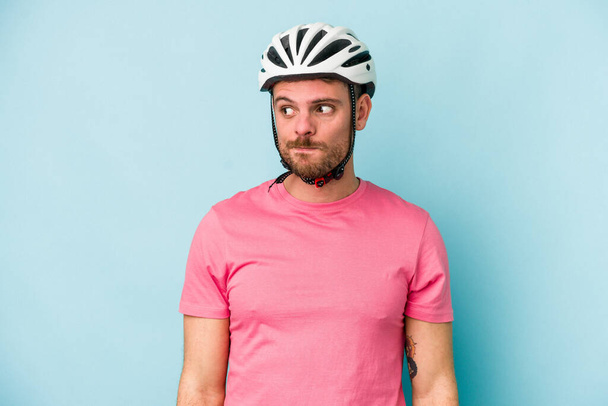 Jonge blanke man met fietshelm geïsoleerd op blauwe achtergrond verward, voelt zich twijfelachtig en onzeker. - Foto, afbeelding