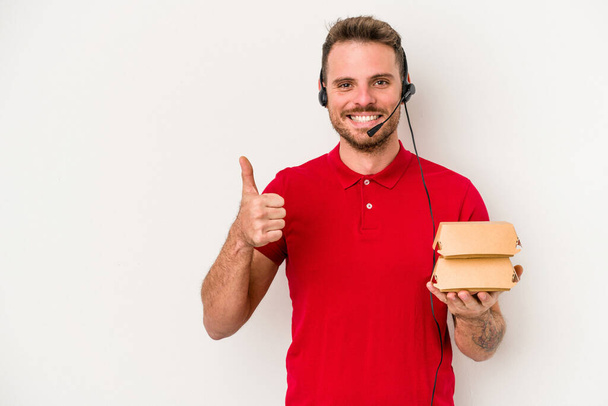 Young caucasian delivery man holding a burger isolated on white background smiling and raising thumb up - Φωτογραφία, εικόνα