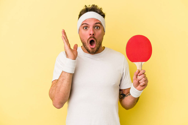 Jonge blanke man met ping pong racket geïsoleerd op gele achtergrond verrast en geschokt. - Foto, afbeelding