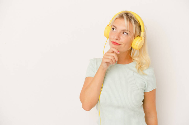 Jonge Russische vrouw luisteren naar muziek geïsoleerd op witte achtergrond kijken zijwaarts met twijfelachtige en sceptische uitdrukking. - Foto, afbeelding