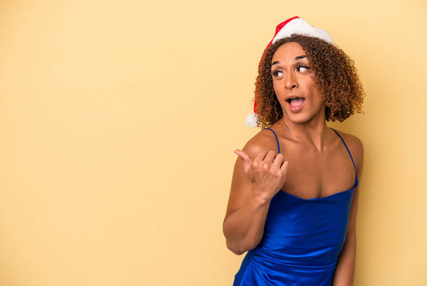 Young latin transsexual woman celebrating christmas isolated on yellow background points with thumb finger away, laughing and carefree. - Photo, Image