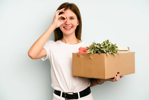 Mladá anglická žena dělá krok při vyzvednutí krabice plné věcí izolovaných na modrém pozadí vzrušený udržet ok gesto na oku. - Fotografie, Obrázek