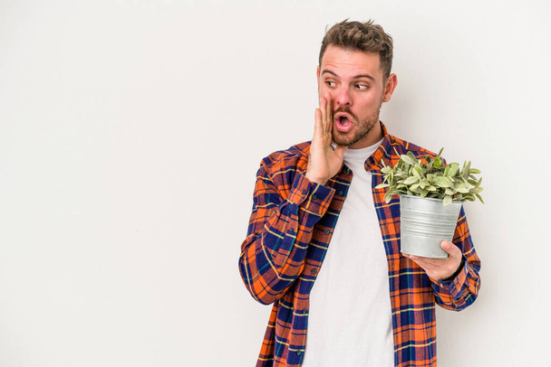Young caucasian man holding a plant isolated on white background is saying a secret hot braking news and looking aside - Foto, imagen