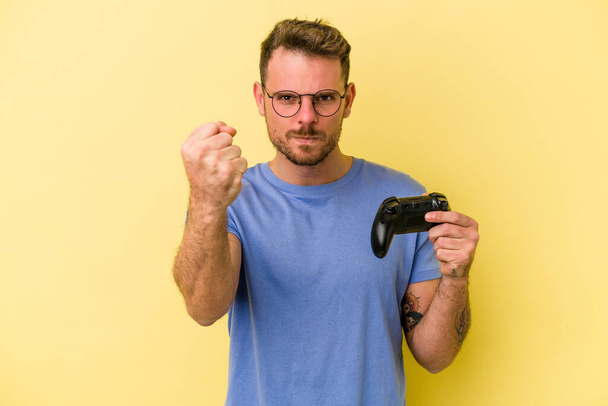 Молодий білий чоловік тримає ігровий контролер ізольований на жовтому тлі, показуючи кулак на камеру, агресивний вираз обличчя
. - Фото, зображення