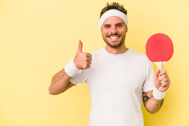 Молодий білий чоловік тримає ракетку для пінг-понг ізольовано на жовтому тлі посміхаючись і піднімаючи великий палець вгору
 - Фото, зображення