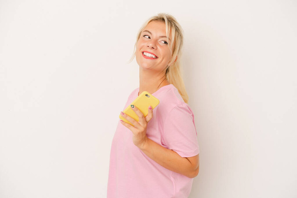 Young Russian woman holding mobile phone isolated on white background looks aside smiling, cheerful and pleasant. - 写真・画像