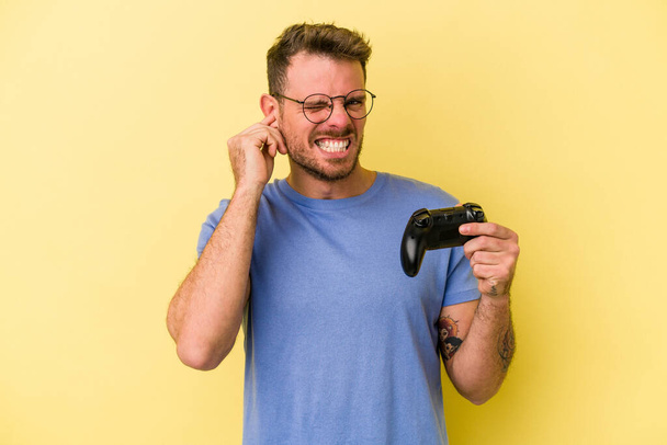 Giovane uomo caucasico che tiene un controller di gioco isolato su sfondo giallo che copre le orecchie con le mani. - Foto, immagini