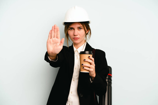 Young architect English woman with helmet and holding blueprints isolated on blue background standing with outstretched hand showing stop sign, preventing you. - Photo, Image