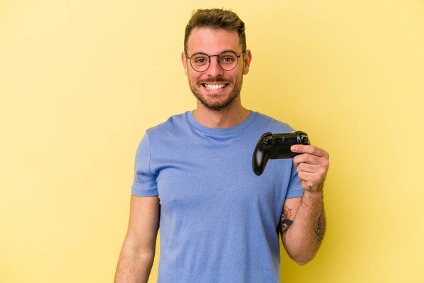młody biały mężczyzna trzyma a gra kontroler odizolowany na żółty tło szczęśliwy, uśmiechnięty i wesoły. - Zdjęcie, obraz