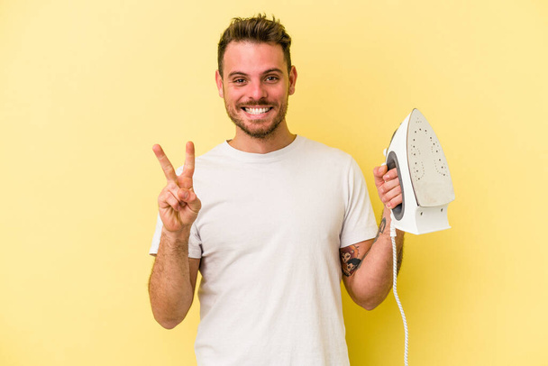 Young caucasian man holding a iron isolated on yellow background showing number two with fingers. - Fotografie, Obrázek