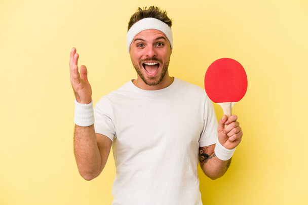 Young caucasian man holding ping pong racket isolated on yellow background receiving a pleasant surprise, excited and raising hands. - Photo, Image