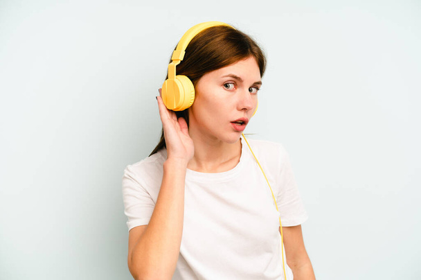 Młoda Angielka słuchająca muzyki odizolowanej na niebieskim tle próbująca słuchać plotek. - Zdjęcie, obraz