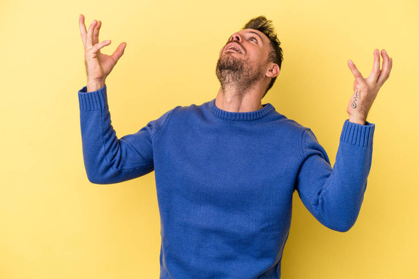Young caucasian man isolated on yellow background screaming to the sky, looking up, frustrated. - Photo, Image