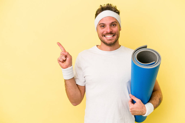 Young sport caucasian man going to yoga classes while holding a mat isolated on yellow background smiling and pointing aside, showing something at blank space. - Foto, afbeelding