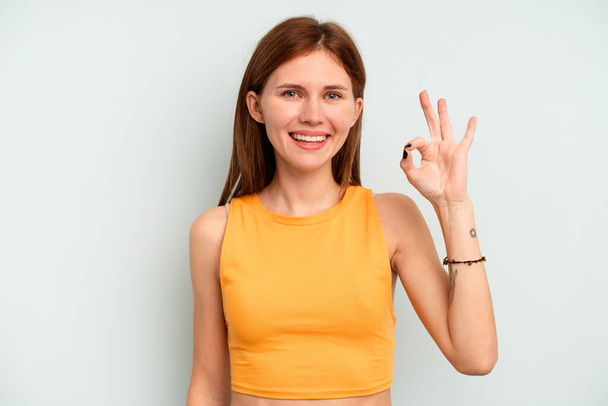 Young English woman isolated on blue background winks an eye and holds an okay gesture with hand. - Photo, Image