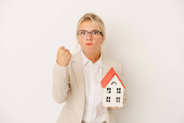 Junge Immobilienmaklerin mit einem Hausmodell auf weißem Hintergrund, das die Faust in die Kamera zeigt, aggressiver Gesichtsausdruck. - Foto, Bild
