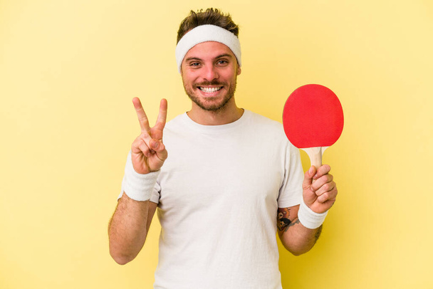 Jeune homme caucasien tenant raquette ping pong isolé sur fond jaune montrant numéro deux avec les doigts. - Photo, image