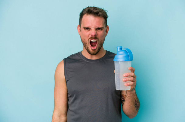 Fiatal fehér férfi iszik tejturmix elszigetelt kék háttér sikoltozó nagyon dühös és agresszív. - Fotó, kép