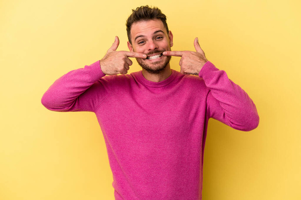 Jeune homme caucasien isolé sur fond jaune sourit, pointant les doigts vers la bouche. - Photo, image