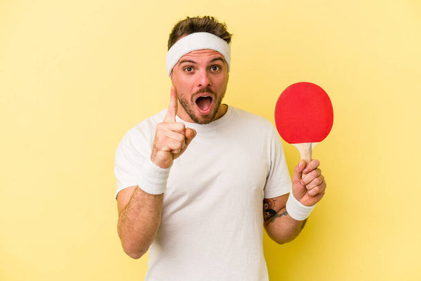 Young caucasian man holding ping pong racket isolated on yellow background having an idea, inspiration concept. - Photo, Image