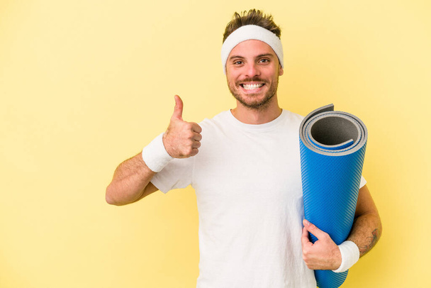 Jovem esporte caucasiano homem indo para aulas de ioga enquanto segurando um tapete isolado no fundo amarelo sorrindo e levantando o polegar para cima - Foto, Imagem