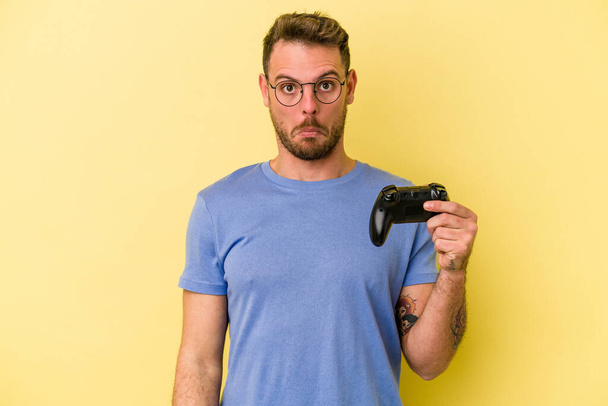 Young caucasian man holding a game controller isolated on yellow background shrugs shoulders and open eyes confused. - Photo, Image
