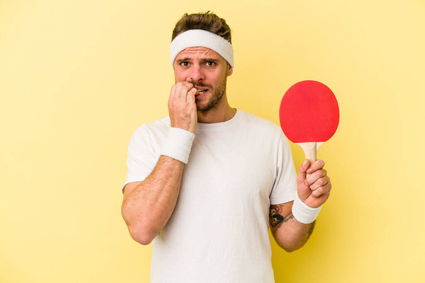 Beyaz tenli genç adam masa tenisi raketi tutuyor sarı arka planda izole edilmiş tırnak yiyor, gergin ve endişeli.. - Fotoğraf, Görsel