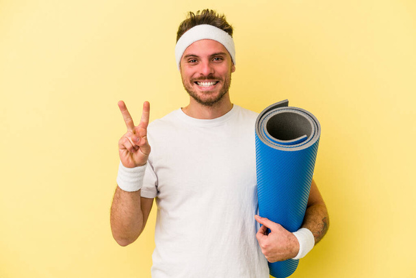 Young sport caucasian man going to yoga classes while holding a mat isolated on yellow background showing number two with fingers. - Photo, Image