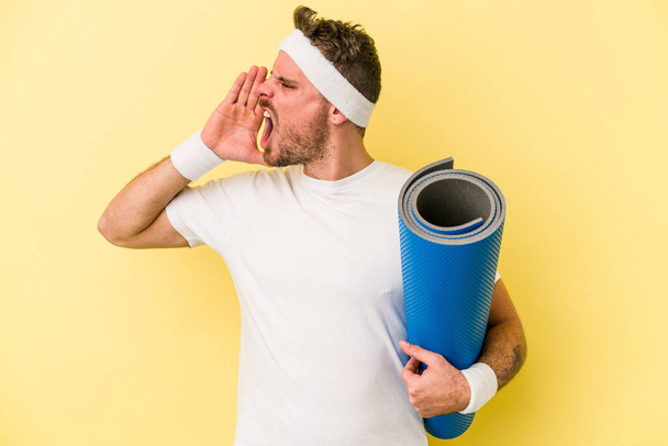 Young sport caucasian man going to yoga classes while holding a mat isolated on yellow background shouting and holding palm near opened mouth. - Foto, afbeelding