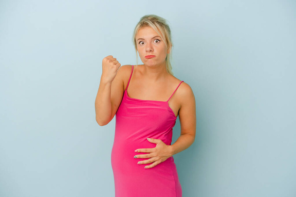 Молода російська жінка, вагітна на синьому фоні, показує кулак камери, агресивний вираз обличчя.. - Фото, зображення