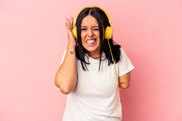 Nuori valkoihoinen nainen, jolla on yksi käsi, kuuntelemassa musiikkia, joka on eristetty vaaleanpunaisella taustalla peittäen korvat käsillään. - Valokuva, kuva