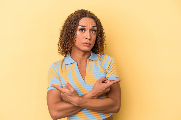 Junge lateinamerikanische transsexuelle Frau isoliert auf gelbem Hintergrund zeigt seitwärts, versucht, zwischen zwei Optionen zu wählen. - Foto, Bild