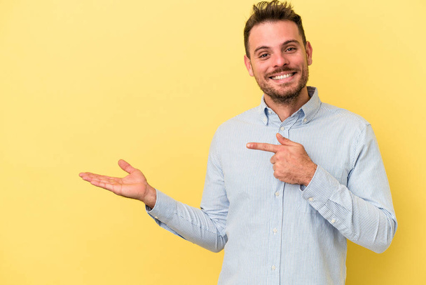 Young caucasian man isolated on yellow background excited holding a copy space on palm. - Photo, Image