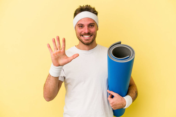 Junger Sportkaukasier geht zu Yoga-Kursen und hält eine Matte isoliert auf gelbem Hintergrund, lächelt fröhlich und zeigt Nummer fünf mit den Fingern. - Foto, Bild