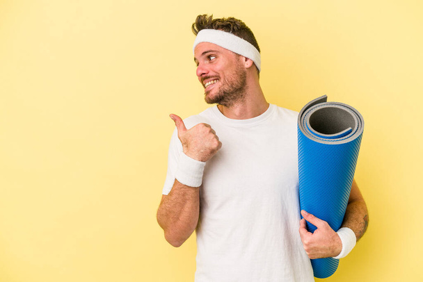 Junger Sportkaukasier geht zu Yoga-Kursen, während er eine Matte isoliert auf gelbem Hintergrund hält, Daumen weg, lachend und unbeschwert. - Foto, Bild