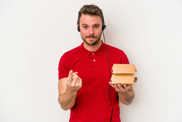 Beyaz arka planda izole edilmiş bir hamburger tutan beyaz tenli genç teslimatçı sanki seni davet ediyormuş gibi parmakla işaret ediyor.. - Fotoğraf, Görsel