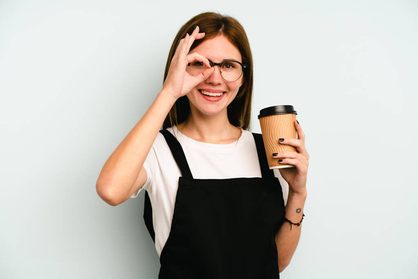 Restauracja kelnerka kobieta trzyma zabrać kawę odizolowany na niebieskim tle podekscytowany utrzymanie ok gest na oko. - Zdjęcie, obraz