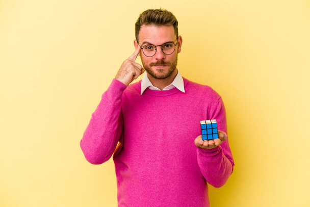 Jeune homme caucasien tenant Rubiks cube isolé sur fond jaune pointant temple avec le doigt, penser, concentré sur une tâche.  - Photo, image