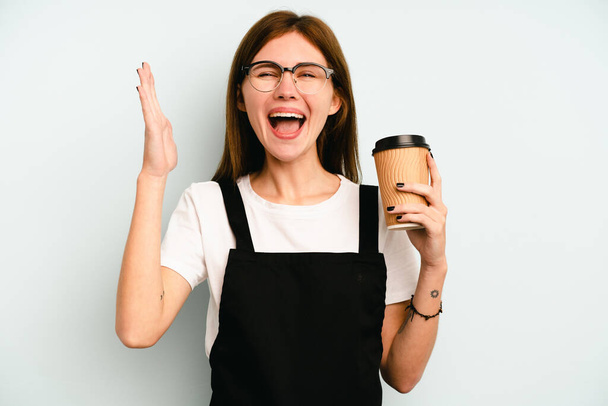 Restaurante garçom mulher segurando um café take away isolado no fundo azul recebendo uma agradável surpresa, animado e levantando as mãos. - Foto, Imagem