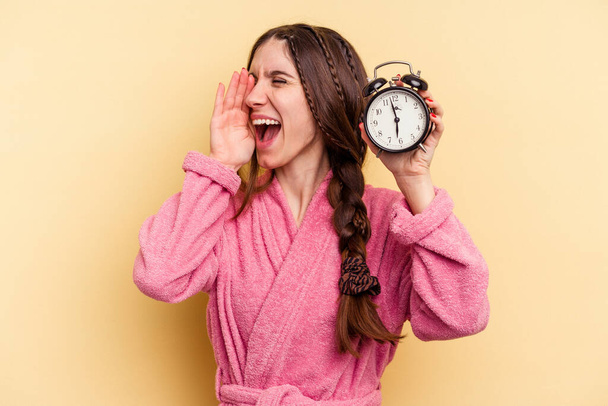 Mujer caucásica joven con un albornoz sosteniendo un reloj despertador aislado sobre fondo amarillo gritando y sosteniendo la palma cerca de la boca abierta. - Foto, Imagen